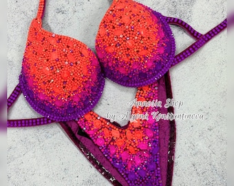 May include: A purple and orange ombre bikini competition suit with crystal embellishments. The suit features a halter top and a high-cut bottom. The text "Amnesia Shop by Alyona Konstantinova" is printed on the suit.
