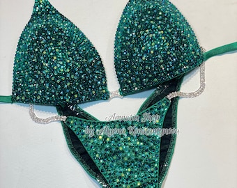May include: Green sparkly bikini competition suit with crystal embellishments. The suit has a halter top and a high-cut bottom. The text "Amnesia Shop by Alyona Konstantynova" is printed on the bottom of the suit.