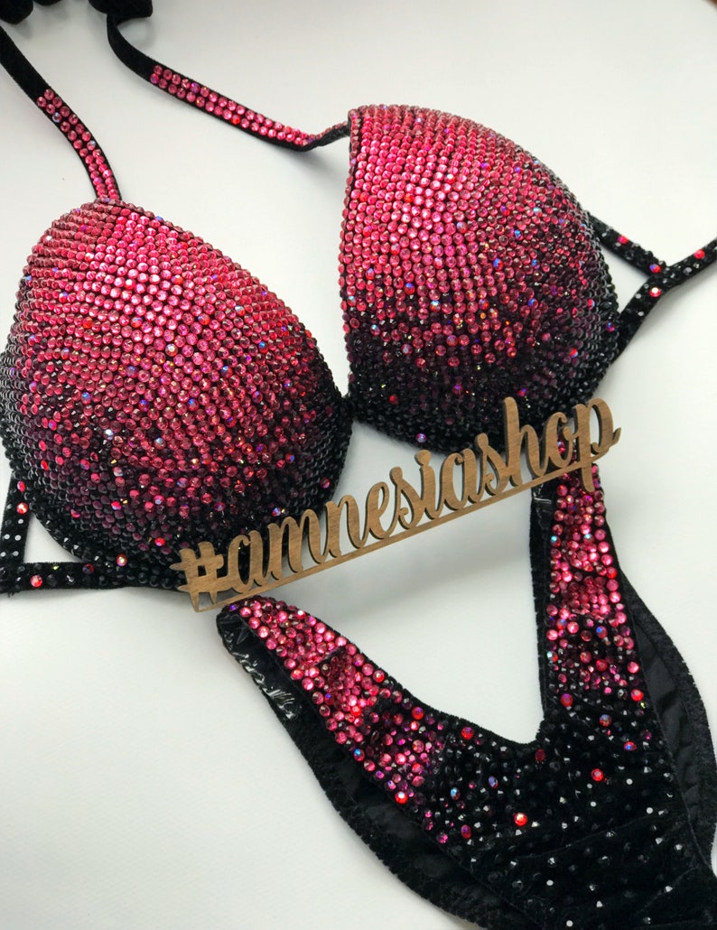 May include: A black and pink competition bikini with a gradient of rhinestones. The bikini has a V-shaped neckline and a high-cut leg. The bikini is decorated with rhinestones in a gradient from black to pink. The bikini is also decorated with a black velvet trim. The bikini is on a white background. #annesiashp