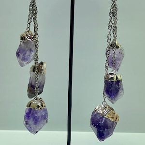 Può includere: Un paio di orecchini pendenti con cristalli di ametista grezzi, cappucci in metallo color argento e dettagli a catena. Gli orecchini sono esposti su un supporto nero su sfondo bianco. I cristalli di ametista hanno una tonalità viola intenso.