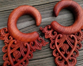 May include: A pair of wooden ear gauges with intricate carved designs. The gauges are a reddish-brown color and have a heart-shaped design in the center.