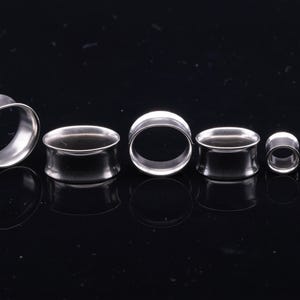 May include: A set of six silver-toned metal ear tunnels arranged in a row, varying in size from largest to smallest. Each piece features a smooth, polished cylindrical shape with flared edges, designed for ear stretching and body modification.