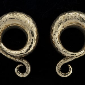 May include: A pair of gold-toned metal ear gauges with a hammered finish and a twisted design. The gauges have a circular opening and a long, curved tail.