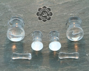 May include: Set of six clear glass ear gauges, including two pairs of plugs and two single flares. The plugs are cylindrical and the flares are slightly curved. The gauges are arranged on a gray stone surface.
