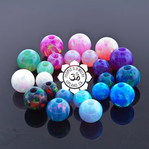 Könnte beinhalten: Sortiment an bunten Glasperlen mit verschiedenen Mustern und Farben, darunter rosa, blau, grün, lila und weiß. Die Perlen sind auf einer schwarzen Oberfläche angeordnet. Die Perlen sind mit "People's Jewelry" mit einem Om-Symbol und "Upland, CA" beschriftet.