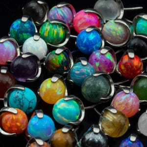 May include: A collection of colorful, round gemstone pendants with silver-tone metal settings. The gemstones display a variety of colors, including blue, green, pink, and orange. The pendants are arranged closely together against a dark background.