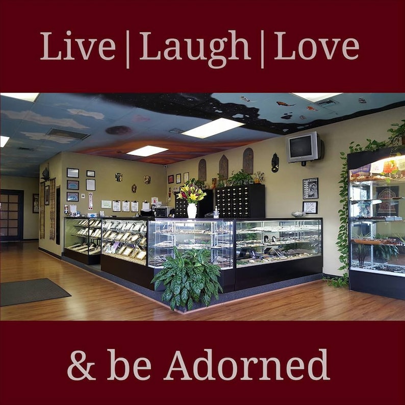 May include: The interior of a retail store featuring glass display cases filled with jewellery and accessories. The store has a wooden floor and a painted ceiling with clouds and stars. The sign above the display cases reads "Live | Laugh | Love & be Adorned".