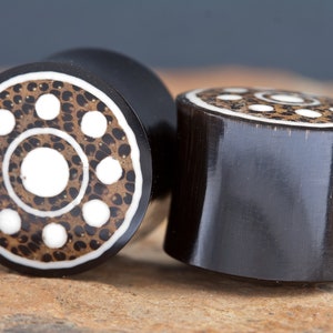 May include: Two black and white wooden ear gauges with a circular pattern. The gauges have a white centre circle surrounded by a ring of smaller white circles.