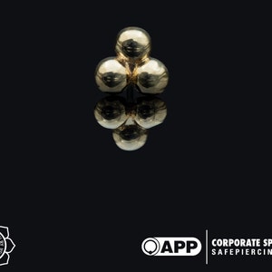 Pode incluir: Um piercing para o corpo de três bolas em ouro. As bolas são lisas e redondas.