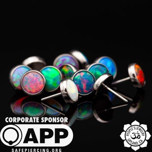 May include: A close-up of a group of colorful opal body jewelry. The jewelry is made of surgical steel and features a variety of opal colors, including blue, green, red, and white. The jewelry is arranged on a black background.