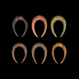 May include: Six wooden ear gauges in different shades of brown, each shaped like a crescent moon.