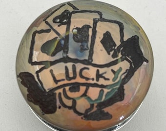以下が含まれることがあります： 銀色の枠にセットされた丸いガラスのcabochonには、トランプと「LUCKY」と書かれたバナーが手描きで描かれています。デザインは、マルチカラーの背景に黒いインクで描かれています。