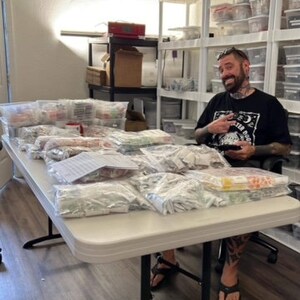 May include: A white table with many clear plastic bags filled with small items. A person is sitting at the table wearing a black t-shirt with a graphic print and black trousers.
