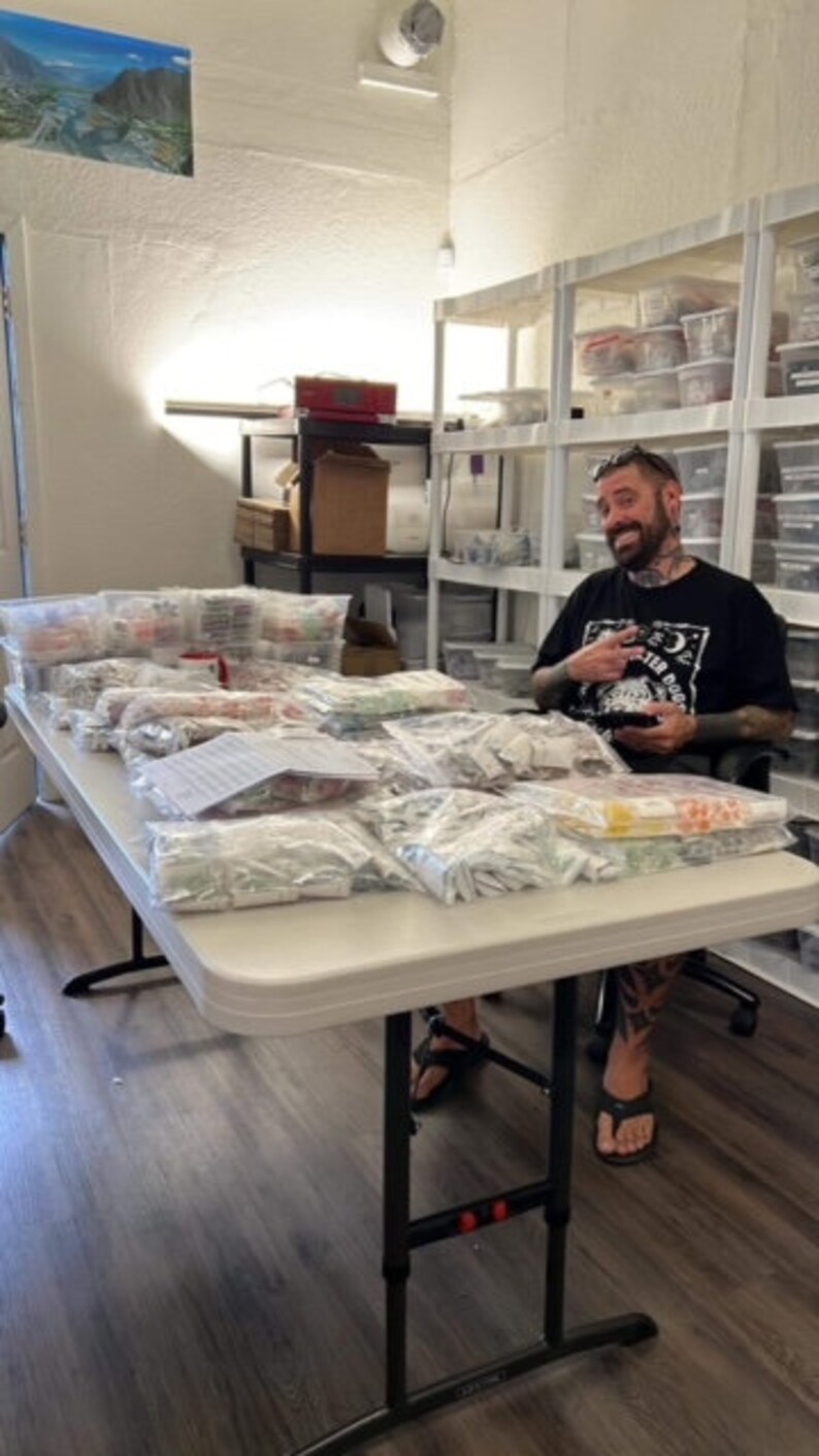 May include: A white table with many clear plastic bags filled with small items. A person is sitting at the table wearing a black t-shirt with a graphic print and black trousers.