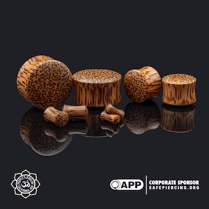 May include: A set of six wooden ear gauges in various sizes. The gauges are made of natural wood with a brown and black speckled pattern. The gauges are arranged in a row on a black surface.