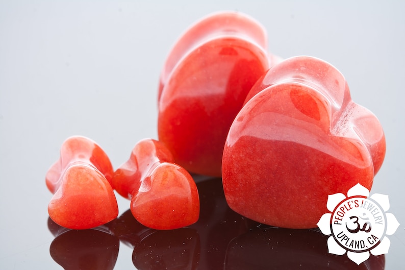 May include: Four heart-shaped red carnelian stones of varying sizes. The largest heart is in the centre of the image, with two smaller hearts to the left and one smaller heart to the right. The hearts are smooth and polished.