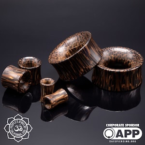 May include: A set of five wooden ear gauges in various sizes. The gauges are made of dark brown wood with a natural grain pattern. The gauges are arranged on a black surface.