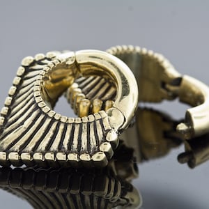 May include: A pair of gold-colored metal ear gauges with a geometric design. The gauges are in the shape of a square with a rounded edge and have a decorative pattern of lines and ridges.