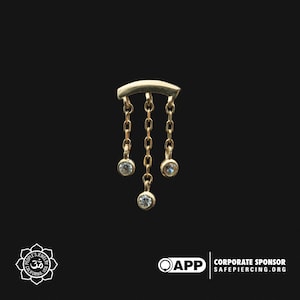 Puede incluir: Pendiente de aro dorado con tres pequeñas piedras preciosas transparentes que cuelgan de una barra curva.