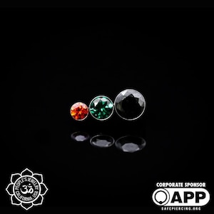 May include: Three small round gems in a row. The gem on the left is orange, the middle gem is green, and the gem on the right is black.