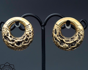 Puede incluir: Un par de expansores de oreja de metal de color dorado con una superficie texturizada. Los expansores se muestran en un soporte negro.