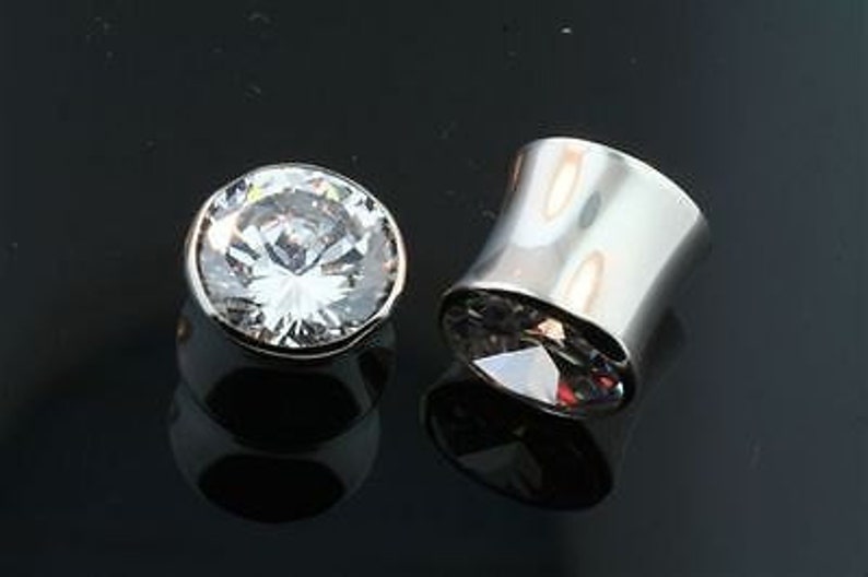 May include: Two silver ear gauges with clear crystal stones. The gauge on the left is a single stone, while the gauge on the right is a flared tube with a stone at the bottom.