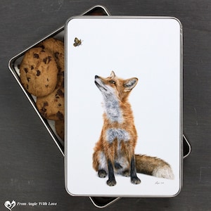 May include: A white metal tin featuring an illustration of a red fox gazing upwards at a butterfly. The fox is depicted against a white background. The tin is open, revealing chocolate chip cookies inside.