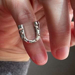 May include: A silver horseshoe ring with small red gemstones set into the metal. The ring is worn on a finger.