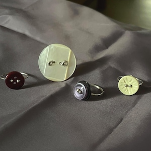 May include: Three rings featuring button tops and one loose circular button. The rings include a dark maroon four-hole button, a purple two-hole button, and a pale yellow patterned button. The loose button is a large, round, iridescent white piece.