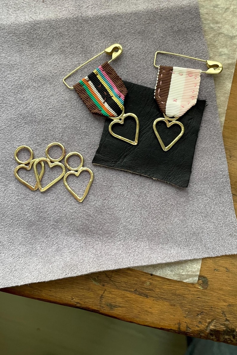 May include: Gold-colored heart-shaped charms and pins with colorful ribbons. Two pins feature a gold heart pendant attached to a ribbon with a safety pin. Three heart charms are also visible.