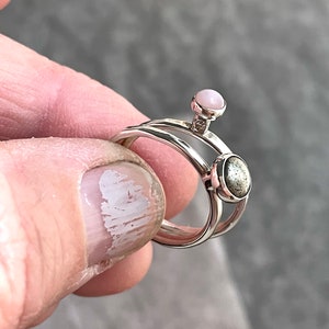 May include: A silver ring set with two stones. The top stone is a light pink oval, and the bottom stone is a dark gray oval.