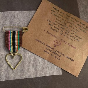 May include: A gold-colored heart-shaped pendant with a colorful striped ribbon, secured by a safety pin. A brown card with text from President Jimmy Carter and the words "LOVE MEDALS" is in the background.