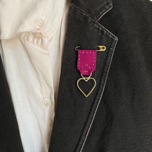 May include: A dark gray blazer with a pink ribbon pin and a gold heart charm. The pin is attached with a gold safety pin. A light-colored button-down shirt is visible underneath the blazer.