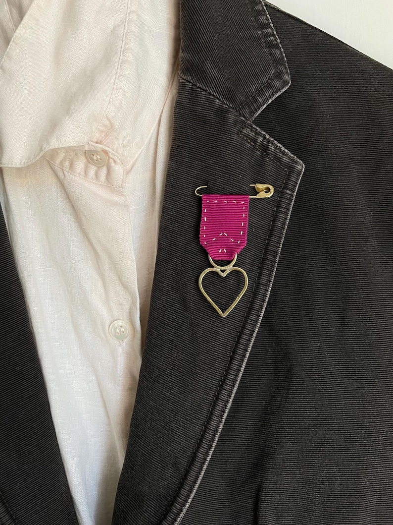 May include: A dark gray blazer with a pink ribbon pin and a gold heart charm. The pin is attached with a gold safety pin. A light-colored button-down shirt is visible underneath the blazer.