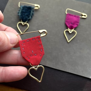 May include: Three decorative brooches with a safety pin design. One brooch has a red ribbon, another has a teal velvet, and the third has a magenta ribbon. Each brooch features a gold-colored heart pendant.