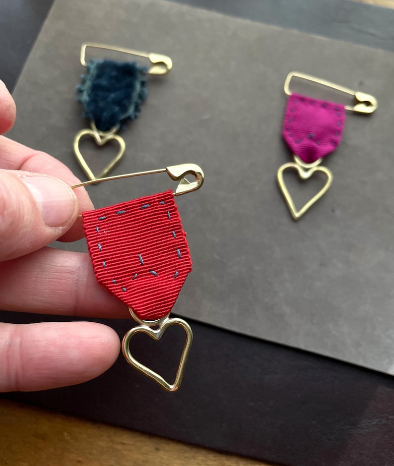 May include: Three decorative brooches with a safety pin design. One brooch has a red ribbon, another has a teal velvet, and the third has a magenta ribbon. Each brooch features a gold-colored heart pendant.