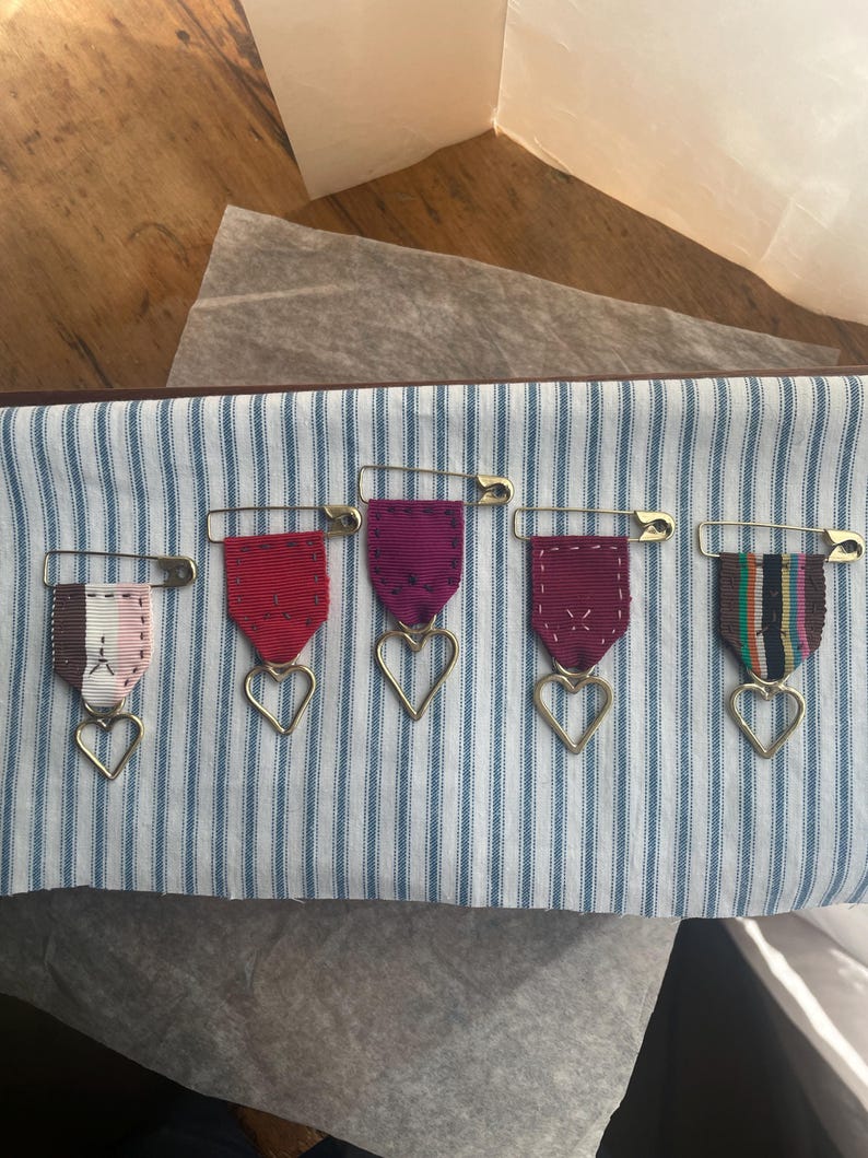 May include: Five decorative ribbon brooches with heart-shaped pendants. Each brooch features a different colored ribbon: pink, red, purple, burgundy, and striped. The brooches are attached with gold safety pins to a blue and white striped fabric.
