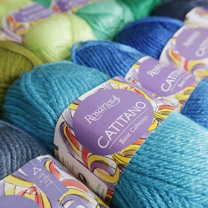 May include: A close-up of several balls of yarn in various colours, including green, blue, and turquoise. The yarn is labelled "Rosarios 4 Catitano Basic Collection".