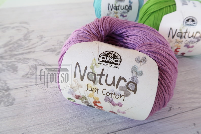 May include: A skein of purple cotton yarn labeled "Natura Just Cotton" by DMC. The yarn is wound around a white label with a floral design.