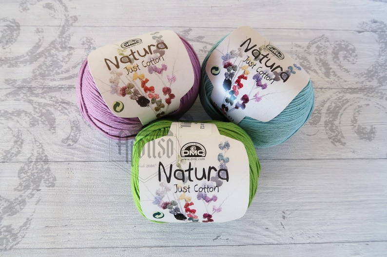 May include: Three balls of yarn in different colors: light purple, green, and light blue. Each ball has a label that says "Natura Just Cotton" with a small flower design.