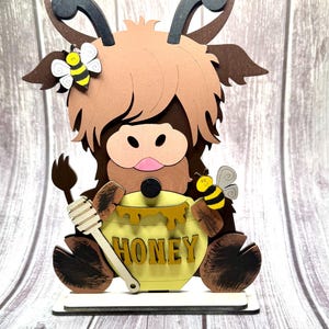 May include: A wooden Highland cow figurine with a honey pot. The cow is brown with a light brown fringe and black horns. A yellow honey pot with the word "HONEY" is held by the cow. Two bees are attached to the cow.