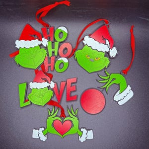 Könnte beinhalten: Satz von sechs Grinch-Weihnachtsanhängern. Die Anhänger sind aus Holz und zeigen das Gesicht des Grinchs, ein rotes Herz, die Worte "Ho Ho Ho" und "Love" sowie einen roten Anhänger.