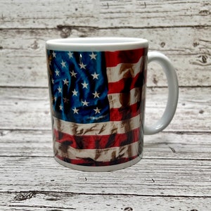 May include: White ceramic mug featuring a detailed American flag design. The mug showcases the flag's blue field with white stars and red and white stripes. The handle is white, and the mug is set against a light wood-grain background.