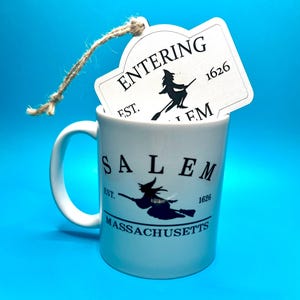 May include: White ceramic mug with a black silhouette of a witch flying on a broomstick. The mug says "Salem, Est. 1626, Massachusetts". A white tag with black text says "Entering Salem, Est. 1626" with a witch silhouette on a broomstick.