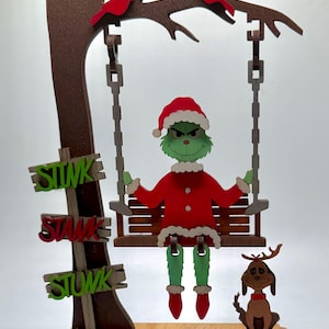 May include: A decorative wooden cutout featuring a green character in a red Santa suit sitting on a swing suspended from a brown tree. The tree has two red birds, a brown dog with antlers, and three signs stacked vertically that each read STINK STANK STUNK.