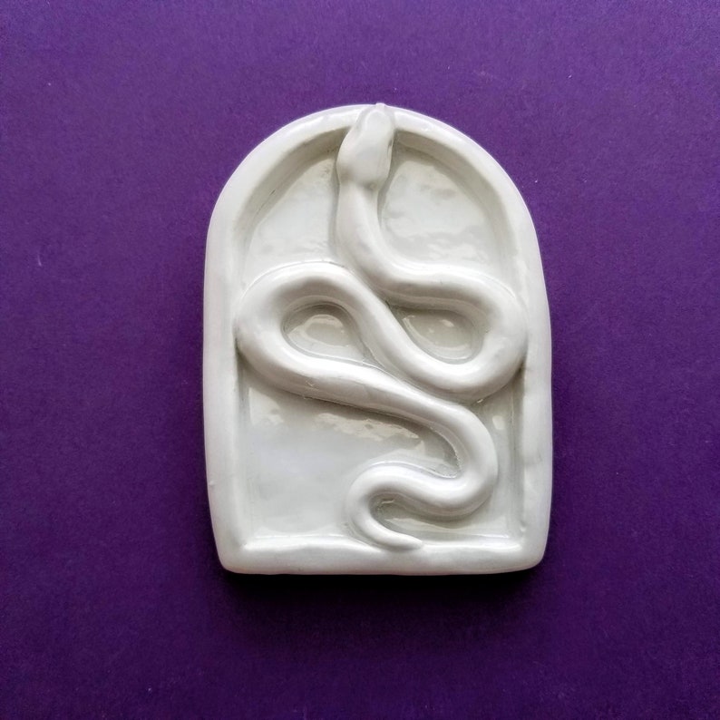 May include: White ceramic wall decor featuring a raised snake design in an arched frame.