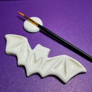 Glossy white bat with wings out spread . each dip in the wing is a little indenture . small circular brush rest and black paint brush at the top . background is deep purple.