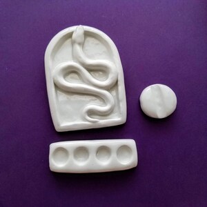 May include: White ceramic paint palette with six round wells and a separate snake-shaped well. The palette is on a purple surface.