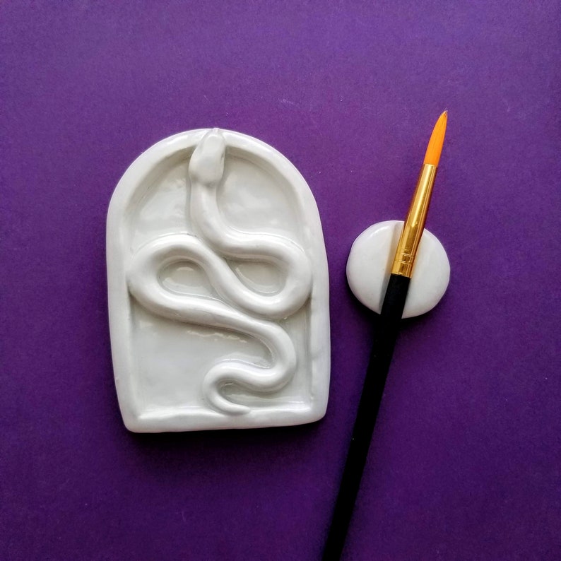 May include: White ceramic dish with a raised snake design. The dish is shaped like an arch and has a smooth, glossy finish. A small white ceramic brush holder is next to the dish. A paintbrush with a black handle and gold bristles is in the brush holder.