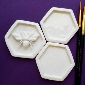 May include: Three white ceramic hexagonal dishes, one with a raised bee design, on a purple background.